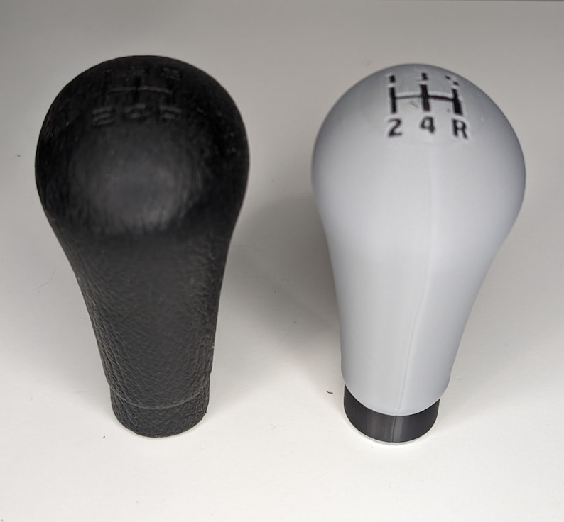 May include: Two gear shift knobs, one black and one white. The black knob has a textured surface and the white knob is smooth. Both knobs have a gear shift pattern on the top.