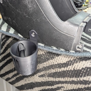May include: A dark grey, 3D-printed cup holder attached to a black surface. The cup holder has a cylindrical shape with a small, curved handle and the numbers "450" printed on the side. The holder is mounted with a single screw.