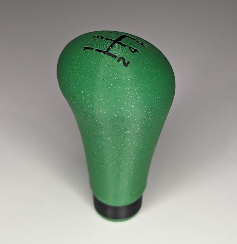 May include: Green gear shift knob with a black 5-speed pattern on top. The knob has a textured surface.