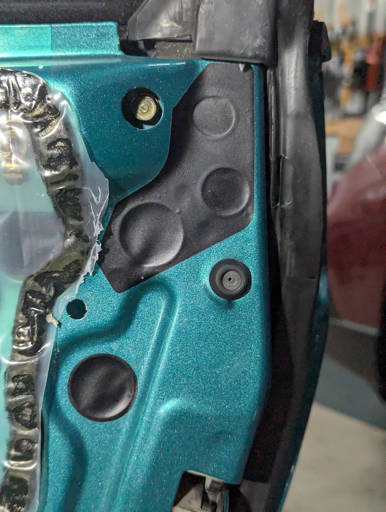 May include: Close-up of a teal car door frame with black plastic components. Visible are a bolt, a rubber grommet, and various circular indentations. The door frame has a metallic finish.