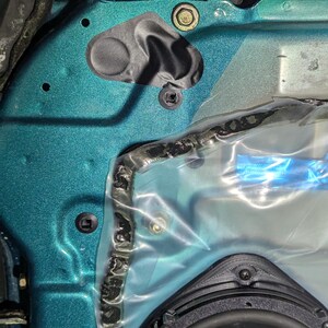 May include: Close-up of a teal car door panel with various components. Visible are a black speaker, plastic sheeting, and several fasteners, including screws and a bolt. The panel has a textured finish.