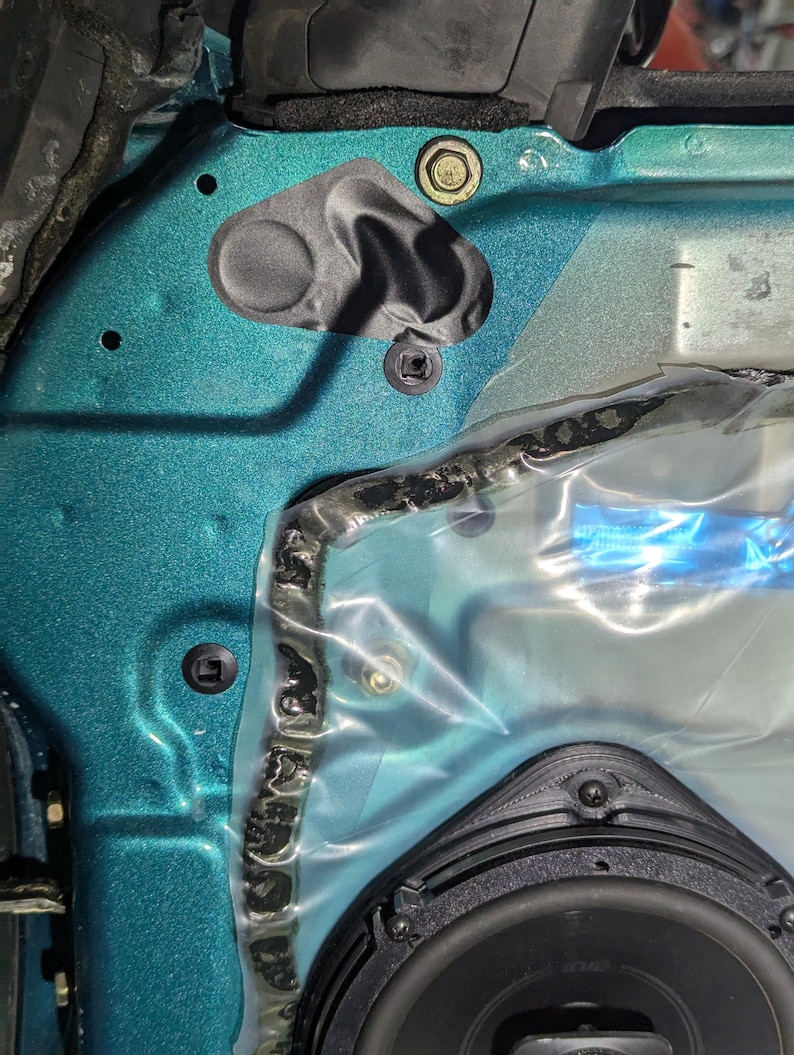 May include: Close-up of a teal car door panel with various components. Visible are a black speaker, plastic sheeting, and several fasteners, including screws and a bolt. The panel has a textured finish.