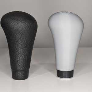 May include: Two gear shift knobs, one black leather and one gray plastic. Both have a similar shape and design.
