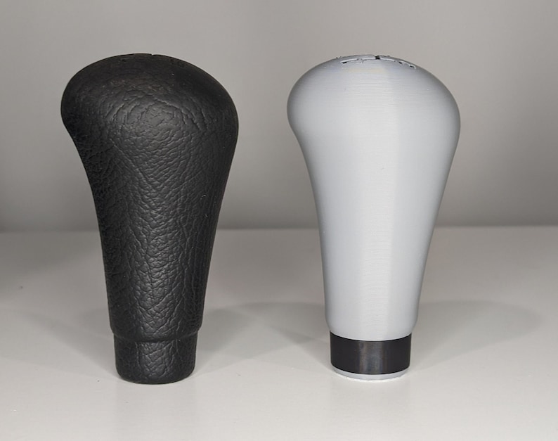 May include: Two gear shift knobs, one black leather and one gray plastic. Both have a similar shape and design.