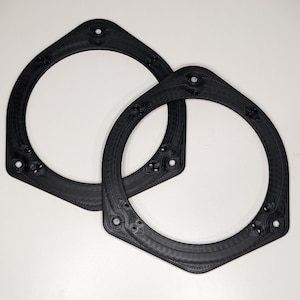 May include: Two black plastic speaker rings, likely for car audio, with a hexagonal shape and multiple small holes.