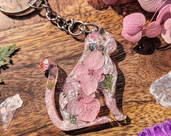 Puede incluir: Un llavero de gato de resina rosa con un llavero plateado. El gato está lleno de flores rosas prensadas y purpurina.