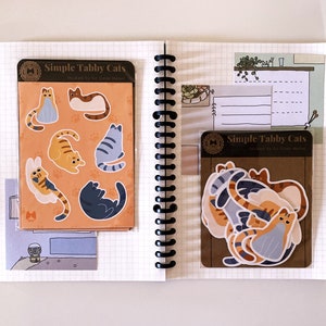 May include: Two sheets of stickers featuring illustrations of tabby cats in various poses. The stickers are packaged in clear plastic sleeves with brown paper backing. The text on the packaging reads "Simple Tabby Cats" and "Stickers by Glass House Market".