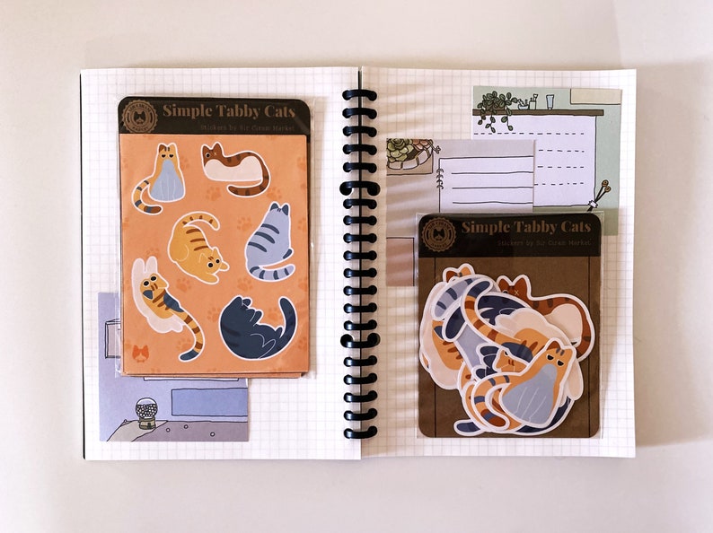 May include: Two sheets of stickers featuring illustrations of tabby cats in various poses. The stickers are packaged in clear plastic sleeves with brown paper backing. The text on the packaging reads "Simple Tabby Cats" and "Stickers by Glass House Market".