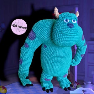 K&ouml;nnte beinhalten: Ein blaues Monster aus H&auml;kelarbeit mit grauen Flecken und einem grinsenden Mund. Das Monster hat zwei H&ouml;rner und steht auf einer wei&szlig;en Oberfl&auml;che.