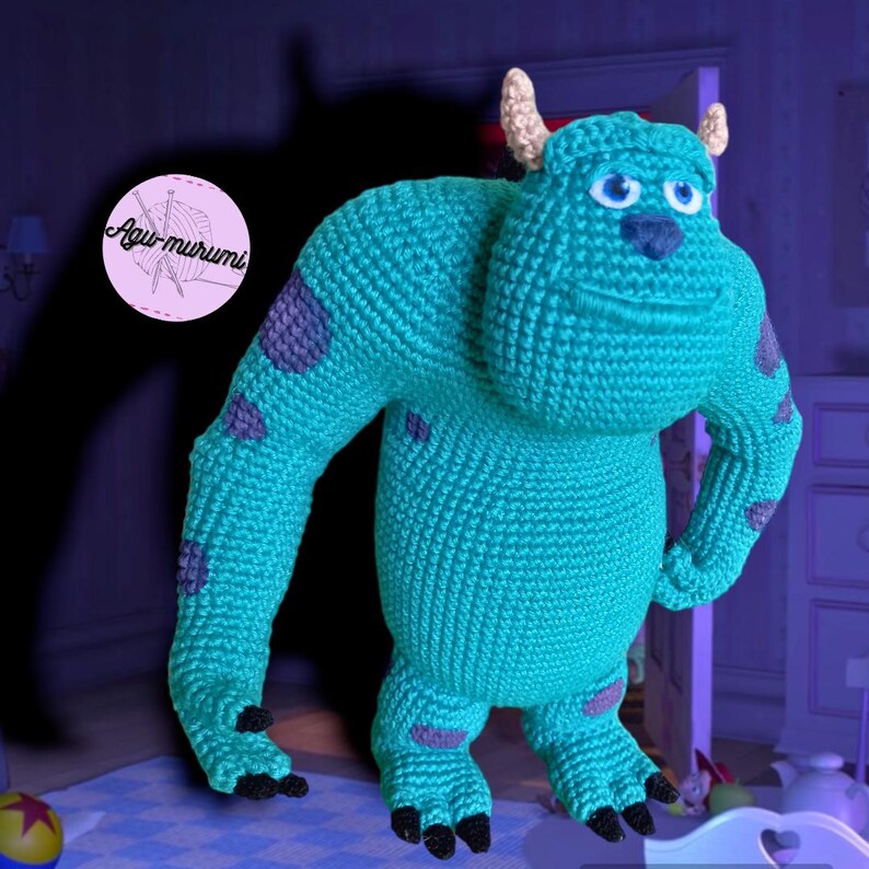 K&ouml;nnte beinhalten: Ein blaues Monster aus H&auml;kelarbeit mit grauen Flecken und einem grinsenden Mund. Das Monster hat zwei H&ouml;rner und steht auf einer wei&szlig;en Oberfl&auml;che.
