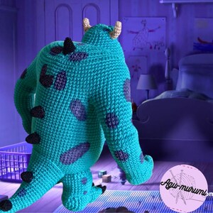 K&ouml;nnte beinhalten: Ein blaues und violettes H&auml;keltier aus Pl&uuml;sch, das ein Monster mit gro&szlig;en F&uuml;&szlig;en und einem einzigen Horn darstellt. Das Spielzeug steht auf einem gestreiften Teppich in einem Kinderzimmer.