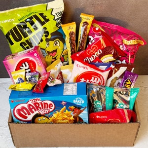 May include: A cardboard box filled with a variety of snack foods, including bags of crisps, chocolate bars, and biscuits. The box is decorated with the words "Marine Boy" and "Baked".