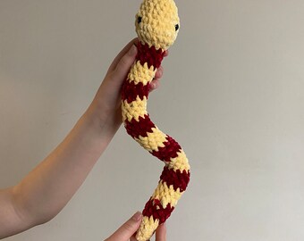 May include: A handmade crochet snake toy in a cream and burgundy color scheme. The snake has a cream-colored head and a body with alternating burgundy and cream stripes. The snake has small black eyes.