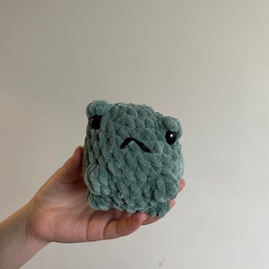 May include: A small, green crocheted plush toy with a grumpy expression. The toy has black eyes and a stitched-on frown.