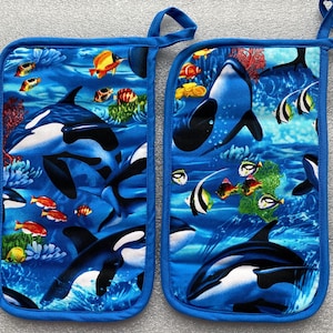 May include: A pair of blue oven mitts with a colorful underwater scene featuring orcas, fish, and coral. The mitts are made of a soft, quilted fabric and have a blue trim.