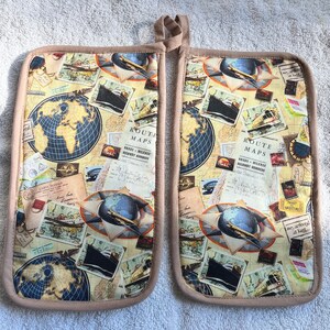 May include: A pair of brown oven mitts with a vintage travel theme. The mitts feature a collage of images including maps, globes, airplanes, and ships. The text "Route Maps" is visible on the mitts.