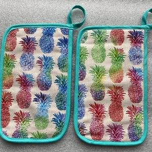 May include: A pair of teal blue oven mitts with a rainbow pineapple print. The mitts are made of a cotton fabric and have a loop for hanging.