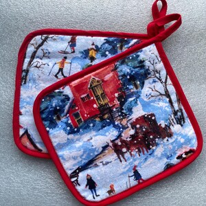 May include: A pair of red and white pot holders with a winter scene design. The design features a red house, snow-covered trees, and people skiing.