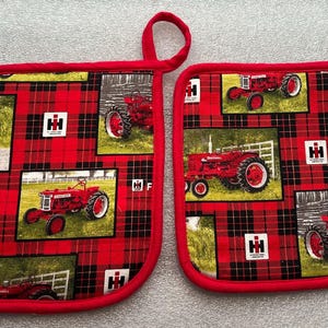 May include: Two red and black plaid oven mitts with red trim and hanging loops. Each mitt features multiple images of red tractors on a green and white background, with the International Harvester logo.
