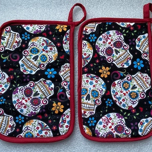 May include: A pair of red and white oven mitts with a black background and a sugar skull pattern. The skulls are white with black outlines and have colorful floral designs.