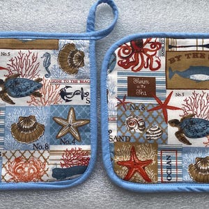 May include: A pair of blue and white pot holders with a nautical theme. The pot holders feature a variety of sea creatures, shells, and anchors. The text "By the Sea" is printed on one of the pot holders.