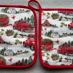 May include: Two red-trimmed pot holders with a winter holiday scene. The design features red pickup trucks carrying wreaths and the text "Happy Holidays." The background includes snow-covered houses, trees, and gifts.