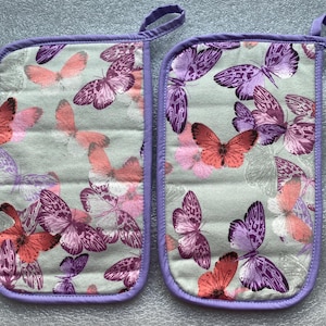 May include: Two purple and white oven mitts with a butterfly pattern. The mitts are made of a soft, quilted fabric and have a loop for hanging.