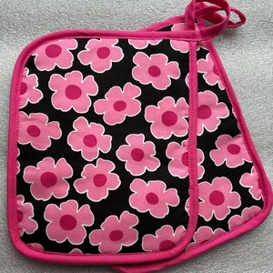 May include: A pair of quilted pot holders with a black background and a pattern of pink flowers. The pot holders are trimmed with a bright pink binding and have a loop for hanging.