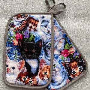 May include: A pair of gray oven mitts with a colorful print of kittens and flowers. The mitts have a loop for hanging.