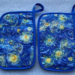 May include: A pair of blue pot holders with a starry night design. The pot holders have a blue border and are made of fabric.