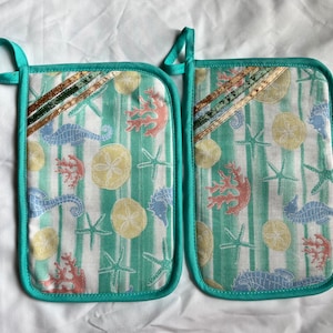 May include: A pair of teal green oven mitts with a white background and a seahorse, starfish, and seashell pattern. The mitts have a brown and gold ribbon trim.