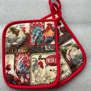 May include: Two red-trimmed pot holders with a rooster and chicken print. The fabric features various rooster illustrations, with text that reads "Fresh Eggs" and "Farmers Market". The pot holders have red loops for hanging.