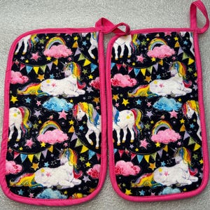 May include: Two oven mitts with a black background featuring a repeating pattern of white unicorns with rainbow manes and tails, rainbows, pink clouds, and stars. The mitts are trimmed with bright pink fabric and have a pink loop for hanging.