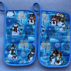 May include: A pair of blue pot holders with a winter snowman theme. The pot holders feature a blue background with white snowflakes and snowmen. The snowmen are wearing scarves and hats.