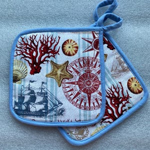 May include: A pair of blue and white pot holders with a nautical theme. The pot holders feature a compass, seashells, starfish, coral, and a ship. The pot holders are trimmed with a light blue fabric.