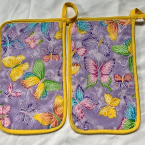 May include: A pair of purple pot holders with a yellow trim and a butterfly pattern. The pot holders are made of fabric and have a glittery finish.