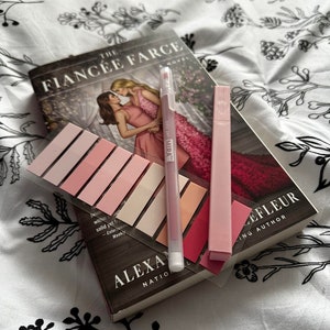 May include: A pink and white book titled "The Fiancée Farce" by Alexa Nation, a bestselling author. The book is on a white bedspread with a black floral pattern. There are pink sticky notes and a pink highlighter on top of the book.