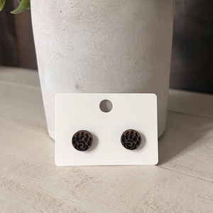 May include: A pair of round, dark brown wooden stud earrings. Each earring features a paw print design. The earrings are displayed on a white earring card, with a grey concrete pot and green plant in the background.