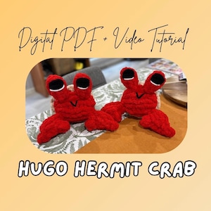 May include: Two handmade red crochet hermit crabs with black eyes and white accents. The crabs are on a table with the text "Digital PDF + Video Tutorial" and "HUGO HERMIT CRAB".