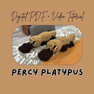 Puede incluir: Dos juguetes de ornitorrinco hechos a mano con ganchillo, cada uno con un cuerpo marrón, pico y pies marrones oscuros y ojos blancos. El texto "Digital PDF + Video Tutorial" y "PERCY PLATYPUS" se muestra sobre un fondo marrón.