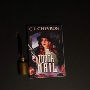 May include: A book titled "TOOTH NAIL" by C.I. Chevron, featuring a woman with a hat and gun on the cover. Beside it is a small, brass-colored object with a dark handle, possibly a letter opener or similar tool.