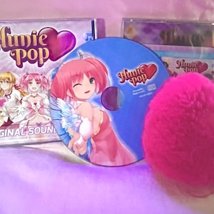 May include: A pink heart-shaped plush toy sits next to a blue CD with the text "Hunie Pop" and a pink heart. The CD is next to a CD case with the text "Hunie Pop Original Sound".