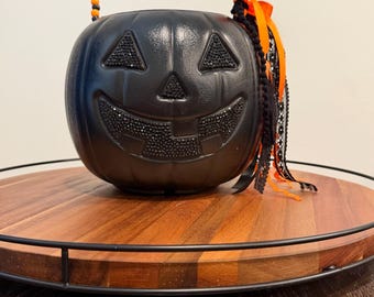 May include: A black plastic pumpkin bucket featuring a jack-o'-lantern face with black rhinestone-encrusted eyes, nose, and mouth. The handle is made of alternating orange and black beads, and a decorative orange ribbon and black lace trim.