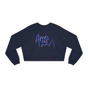 May include: Navy blue cropped sweatshirt with the text "Après Ski" in blue and purple. The sweatshirt has long sleeves and a crew neck. The text is above a mountain graphic.