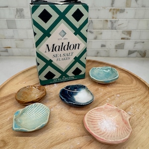 May include: A wooden tray holds six seashell-shaped ceramic dishes in various colors, including blue, pink, and brown. A box of Maldon Sea Salt Flakes is behind the dishes. The box has a green and black diamond pattern.