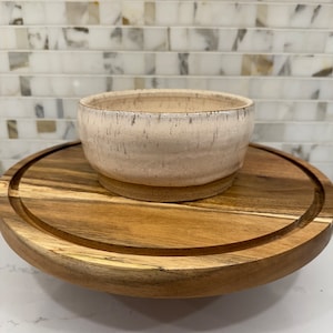 May include: A white ceramic bowl with a speckled glaze sits on a wooden serving tray. The bowl has a rounded shape and a slightly flared rim.