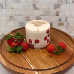 May include: A cylindrical ceramic container with a white base and red cherry branch design. The container sits on a wooden serving board, accompanied by fresh red cherry tomatoes and green parsley.