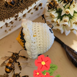 May include: A crocheted bee with black and yellow stripes, wearing a white crocheted hat. The bee is surrounded by white flowers and a few real bees.