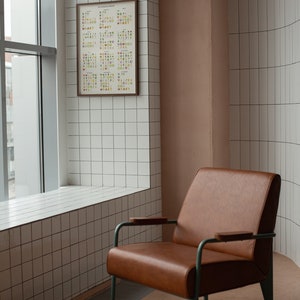 May include: A brown leather armchair with a metal frame and wooden armrests sits in a room with white tiled walls and a window. A framed poster with colorful illustrations is hanging on the wall.