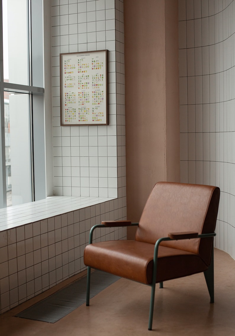 May include: A brown leather armchair with a metal frame and wooden armrests sits in a room with white tiled walls and a window. A framed poster with colorful illustrations is hanging on the wall.